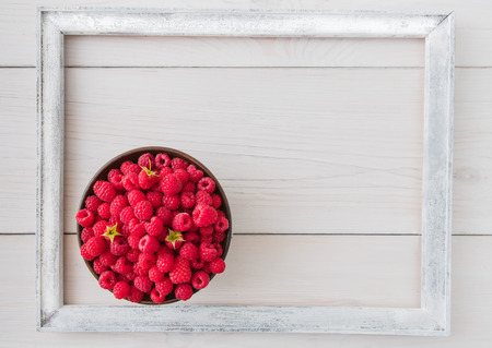 Red fresh raspberries on white rustic wood background. Bowl with natural ripe organic berries with peduncles and white photo frame around it on wooden table, top view with copy spaceの写真素材