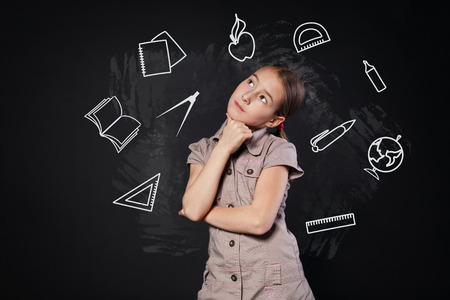 Small girl near blackboard thinking. Thoughtful, pensive child consider something. Smart schoolgirl studio portrait near chalkboard with education icons. Studying school subjects concept.の写真素材