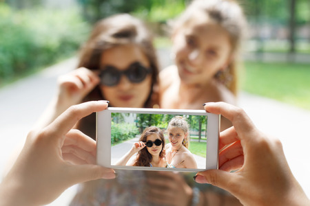 Two girls friends outdoors and someone POV view, taking photos at digital camera of smartphone. Young females at mobile screen, laughing and having fun in summer park. Lifestyle portrait.の写真素材