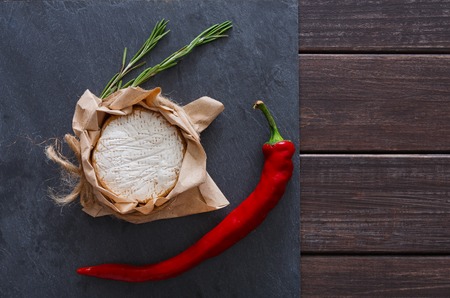 Cheese delikatessen closeup with chili pepper on black stone surface and wood. Camembert or brie circle in brown kraft paper decorated with basil, top view imageの写真素材