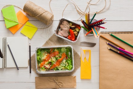 Healthy lunch and diet concept. Take away food in foil boxes, papers and pencils on working table of creative person, designer or student. Salmon and turkey with vegetables, top view on white woodの写真素材
