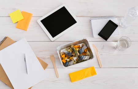 Healthy lunch - foil box with mackerel fish and vegetables with water glass and wooden cutlery on table. Diet food on office table with mobile phone, papers and tablet. Top view, copy spaceの写真素材