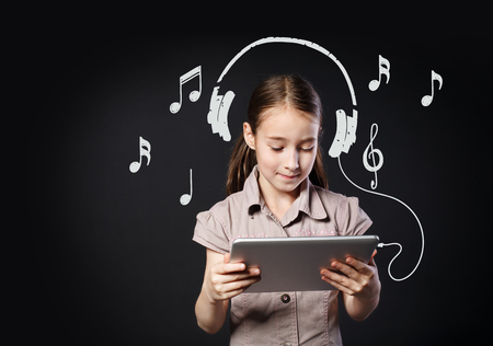 Child and internet music concept. Beautiful cute happy girl with imaginary headphones and musical signs notes, looks at tablet choosing songs to listen, small music lover.の写真素材