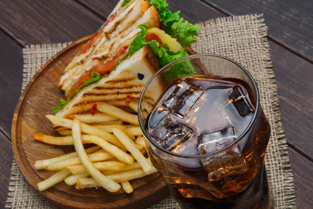 Fast food meals at sandwich bar closeup. Chicken and vegetables sandwich, potato chips and glass of cold cola drink with ice on wood. Take away composition. French fries with snack on wooden desk.の写真素材