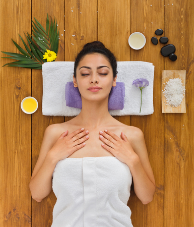 Portrait of woman patient in ayurveda spa wellness center lying relaxed on wood with aroma spa accessories. Young beautiful indian girl with ideal clean skin resting with closed eyes in beauty parlor.の写真素材