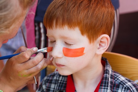 Child animator, artist's hand draws face painting to little boy. Child with funny face painting. Painter makes tiger eyes on boy's face. Children holiday, event, birthday party, entertainment.の写真素材