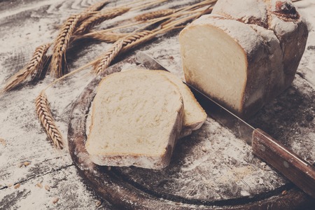 Bakery and grocery concept background. Fresh, healthy toast sliced bread loaf, sprinkled flour, wheat ears, grain and knife cutting it on rustic table and wooden round desk, food closeup.の写真素材