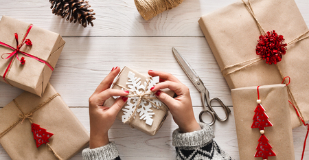Woman's hands wrapping christmas holiday handmade present in craft paper with twine ribbon. Making bow at xmas gift box, decorated with snowflake. Scissors on white wooden table, top view.の写真素材