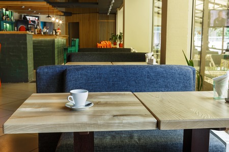 Table with coffee cup in modern restaurant or cafe interior. Public place interior design, bright red wooden chairs and blue sofas, large windows and bar. Morning light, nobody indoors.の写真素材