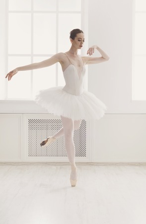 Classical Ballet dancer side view. Beautiful graceful ballerine practice ballet positions in tutu skirt near large window in white light hall. Ballet class training, high-key soft toning.の写真素材