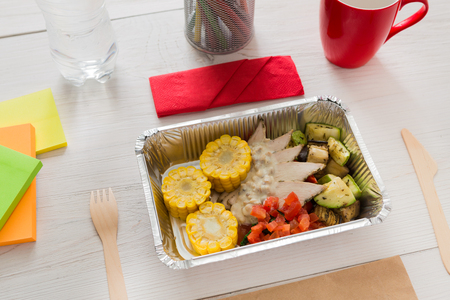 Healthy lunch and diet concept. Take away food in foil boxes, papers and pencils on working table of creative person, designer or student. Meat with corn and vegetables on white woodの写真素材