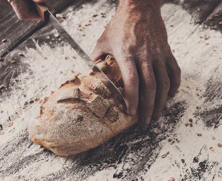 Baking and cooking concept background. Hands of baker closeup cutting bread loaf with knife on rustic wooden table sprinkled with flour. Stained dirty hands of cook.の写真素材