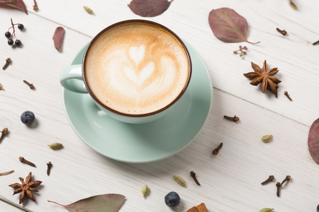 Autumn cappuccino composition. Blue coffee cup with foam, cloves, sloe, dried leaves at white wood background. Fall hot drinks, cafe and bar conceptの写真素材