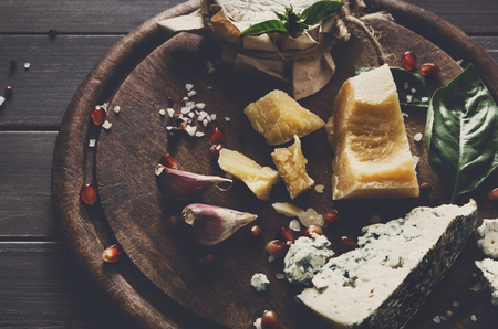 Cheese delikatessen closeup on rustic wood. Wooden round desk with blue cheese roquefort, parmesan, camembert and brie cuts decorated with garlic, pomegranate and basil, french cuisine ingredientsの写真素材