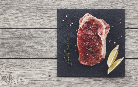 Raw beef steak in craft paper on dark wooden table background, top view. Fresh juicy meat with rosemary and lemon on stone board. Cooking ingredients, butcher's and grocery concept, copy spaceの写真素材