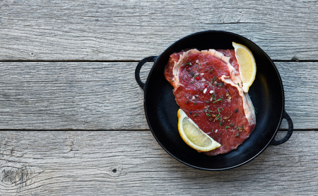 Raw beef steak in craft paper on dark wooden table background, top view. Fresh juicy meat with herbs and lemon in cast iron pan. Cooking ingredients, butcher's and grocery concept, copy spaceの写真素材