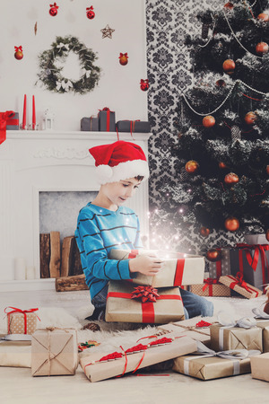 Cute happy boy in santa hat unwrap magic christmas present on holiday morning in beautiful room. Male child open Xmas gift near decorated fir tree and fireplace. Winter holidays conceptの写真素材