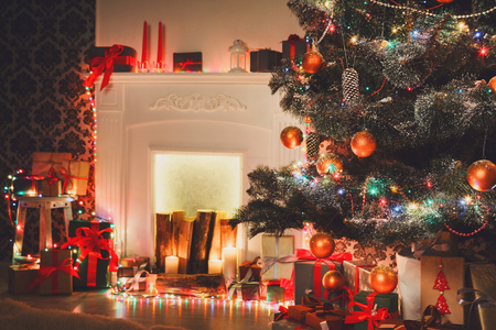 Beautiful holdiay decorated Christmas tree near fireplace, closeup in modern living room. Shining lights, balls and garland on fir tree. Winter holiday night magic. Design and decorations backgroundの写真素材