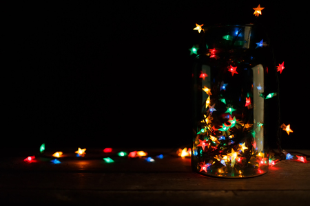Christmas star lights on black background. Holiday shiny garland border spread from glass jar on brown wooden table. Xmas tree decorations, winter holidays illuminationの写真素材
