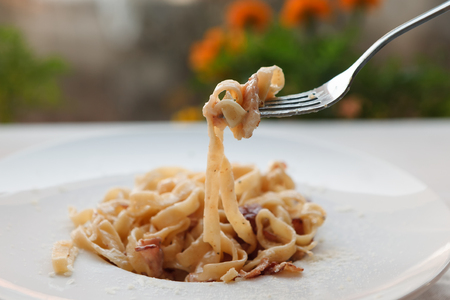 Eating pasta. Fork closeup. Traditional italian carbonara with bacon, egg and parmesan decorated with basil on white round plate at wooden table. Restaurant food, selective focusの写真素材
