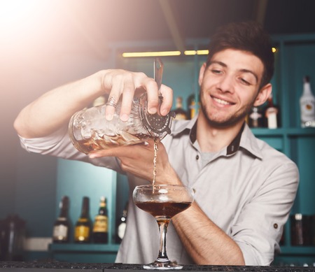 Young handsome Barman in bar interior mixing alcohol cocktail. Professional bartender at work in bar pouring sweet drink into glass on party at night clubの写真素材