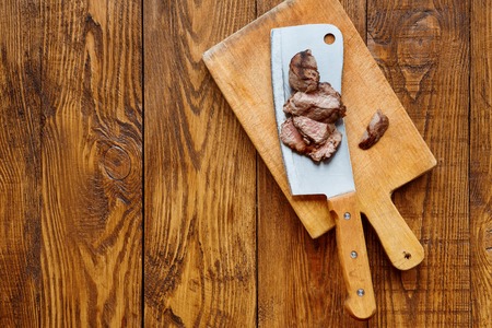 Cleaver butcher's knife slicing grilled steak on meat cutting board on wooden background, top view on copy space. Fresh juicy roasted meat cutsの写真素材