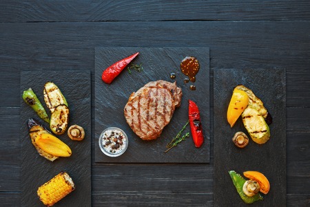 Grilled beef steak and roasted vegetables on dark wooden table background, top view. Juicy meat with rosemary, peppers and cutlery on stone board. Restaurant foodの写真素材