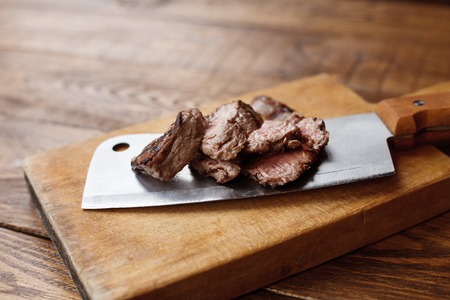 Cleaver butcher's knife slicing grilled steak on meat cutting board on wooden background. Fresh juicy roasted meat cuts. Chef cookingの写真素材