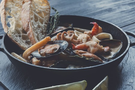Seafood Stew in Saucepan. Authentic italian restaurant cuisine, healthy delicatessen food. Oysters, shrimps, octopus in white cream sauce with bruschetta. Bowl on dark black wood backgroundの写真素材