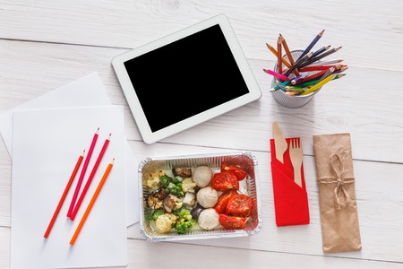 Healthy lunch and diet concept. Take away food in foil box, tablet with copy space and pencils on table of creative person, designer or school student. Meat with vegetables, top view on white woodの写真素材