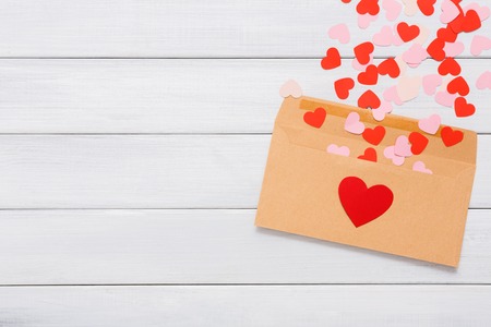 Valentine day love letter. Envelope from craft paper with red hearts heap spread on white wood background. Lovers holiday confession or proposal conceptの写真素材