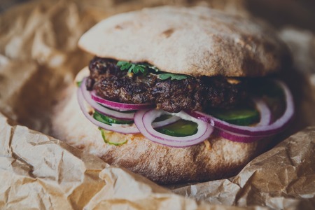 Classic american fast food background. Burger whole grain bun with grilled on barbecue meat and onions on wood in craft paper, closeup. Hamburger with fresh vegetables composition. Filteredの写真素材