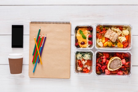 Healthy lunch and diet concept. Take away food in foil box, mobile phone with copy space and pencils on table of artist, designer or student. Meat with vegetables, top view on white woodの写真素材