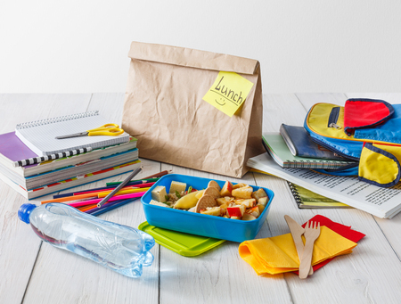 Healthy school lunch for child or teenager. Craft paper package, pile of exercise books, water, bag and food in lunch box on white wood table, cracker with cheese, nuts, oatmeal porridge and applesの写真素材