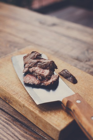 Cleaver butchers knife slicing grilled steak on meat cutting board on wooden background. Fresh juicy...