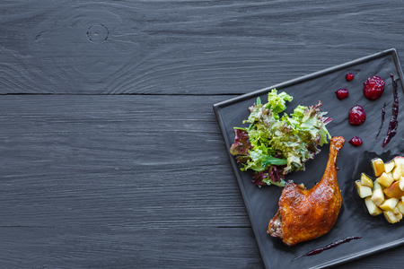 Roasted duck leg closeup served with apples and cherry sauce. Restaurant food on black square slate plate, top view with copy space on wood table backgroundの写真素材