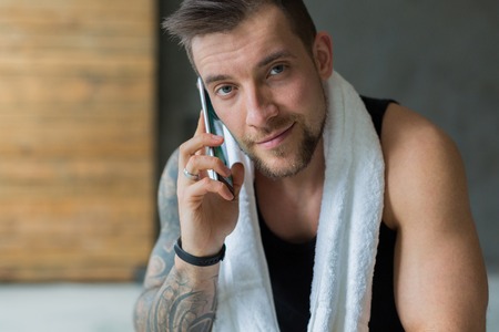 Closeup cropped portrait of handsome athletic man in gym, calling mobile phone. Guy with towel rest after exercise in fitness center. Male model with tattooの写真素材