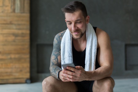 Handsome athletic man in gym, use mobile phone, surfing internet social network. Guy with towel rest after exercise in fitness center. Male model with tattooの写真素材