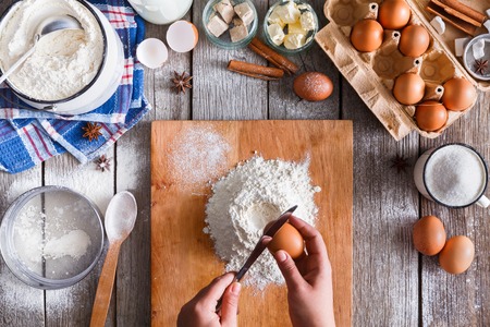 Making dough top view. Overhead of baker hands break egg on flour. Cooking ingredients for pastry on rustic wood, culinary classes or recipe concept.の写真素材