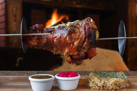 Grill dish in restaurant with fireplace. Grilled roasted pork knuckle closeup on spit with fried sauerkraut cabbage and sauces. Juicy meat meal, german foodの写真素材