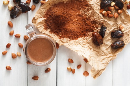 Detox cleanse drink background. Chocolate smoothie ingredients. Natural healthy juice in glass jar for diet or fasting day. Cocoa powder, nuts, date fruit mix on white wood, top viewの写真素材