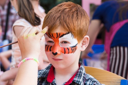 Artists hand draws face painting to little boy. Child with funny face art. Painter makes tiger eyes on boys face. Children holiday, event, birthday party, entertainment.の写真素材