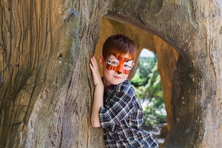 Small boy portrait with funny face art painting. Male child sit in big old tree with tiger drawing on face. Children event, birthday party and creative entertainment.の写真素材