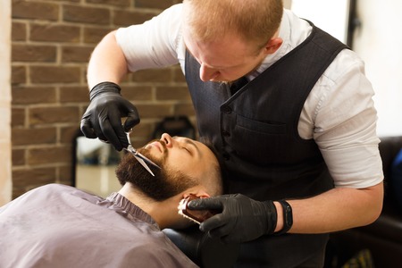 Professional hairdresser wearing black gloves cutting beard of client with scissors at barbershop. Bearded man getting beard haircut at barber salonの写真素材