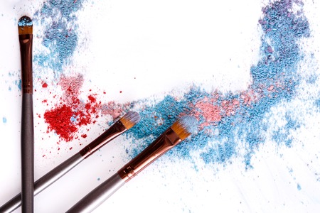 Makeup brushes frame with blush and eyeshadow of pink, blue and coral tones sprinkled on white. Make up and female cosmetics backgroundの写真素材