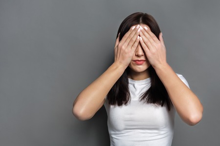 See no evil concept. Portrait of young scared woman covering eyes with hands while standing against gray studio background. Confused girl close eyes with palms ignoring somethingの写真素材