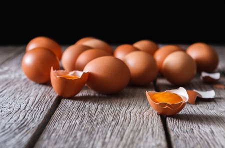 Fresh chicken eggs on wood at black background. Brown and white eggs with cracked eggshell and yellow yolk on rustic table, copy space. Rural still life, natural healthy food concept.の写真素材