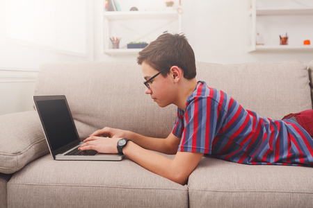 Happy boy with computer on sofa at home. Child engrossed in internet, typing on keyboardの写真素材