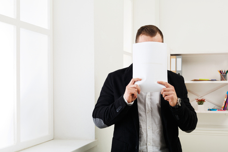 Lot of paperwork. Businessman hiding face with blank paper, standing at office workplace, copy spaceの写真素材