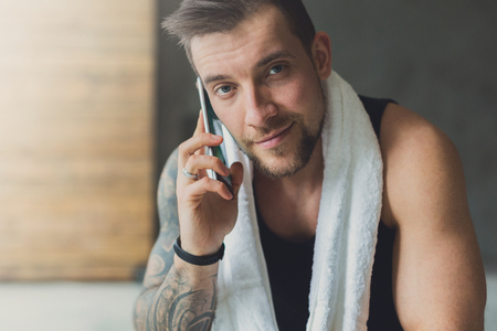 Closeup cropped portrait of handsome athletic man in gym, calling mobile phone. Guy with towel rest after exercise in fitness center. Male model with tattooの写真素材